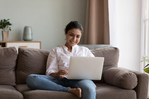Cheerful young Indian freelance employee woman using online app on laptop, working at computer, relaxing on sofa at home, talking on video call, smiling, laughing. Communication concept