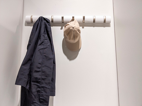 Low angle view of a navy blue jacket and a tan baseball cap hung on a wall hanging rack inside a white, lit up closet
