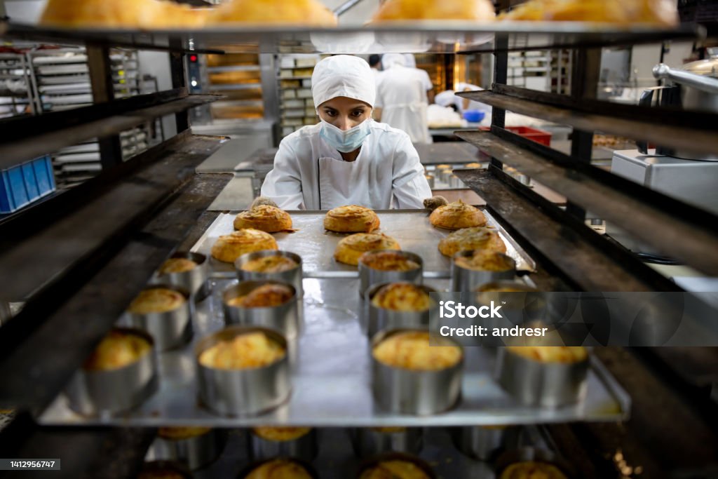 Woman moving a tray of pastries while working at the bakery Latin American woman moving a tray of pastries while working at the bakery - manufacturing concepts Bakery Stock Photo