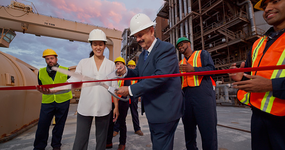 Still shot of an older caucasian man and a young Asian woman wearing suits visiting a power plant for a photo op. They cut a ribbon with a large pair of scissors, while plant workers wearing coveralls, high vis vests and hard hats applaud.