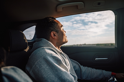 Male authentic portrait of young adult man indoors the car, handsome person smiling and traveling on back seat. Lifestyle photography with happy real people. Blue sky in the window of vehicle. Sunny weather