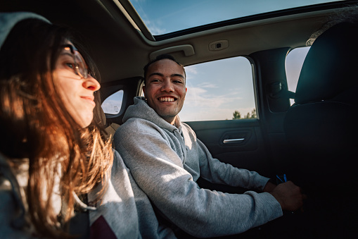 Authentic portrait of young adult people indoors the car, handsome man smiling and traveling on back seat with woman. Lifestyle photography with happy real people. Blue sky in the window of vehicle. Sunny weather