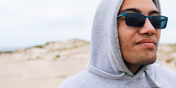 Authentic portrait of young adult man in the sand dunes of desert. Person wear gray sports hoodie and sunglasses, real people walking outdoors. Mixed race guy during the hiking in mountain.