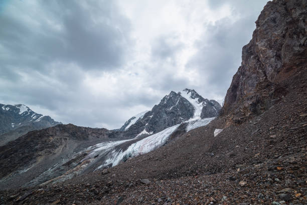 istock Dramatic landscape with hanging glacier against large snow mountain with sharp rocky pinnacle under gray cloudy sky. Glacier with icy cornice in high altitude. Gloomy scenery in mountains in overcast. 1412972141