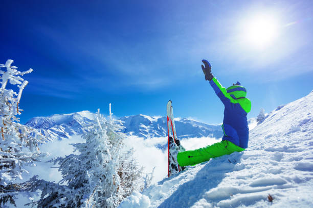 istock Boy with alpine ski sit in snow on Alps mountain top lift hands 1412972732