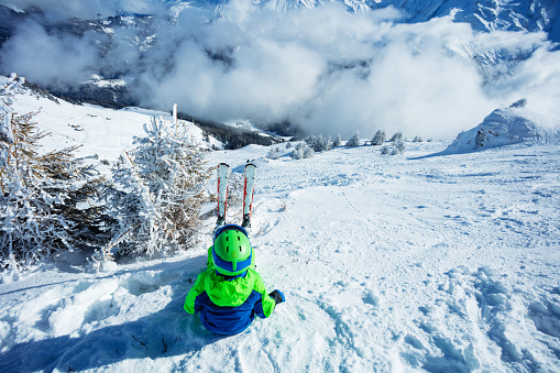 Boy with alpine ski sit in snow lifting hands, wear helmet and sport glasses view from back on top of the mountain over peaks covered by clouds on background