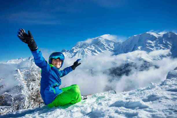 lächelnder junge mit ski alpin und helm sitzt im schnee und hebt die hände - ski skiing european alps resting stock-fotos und bilder