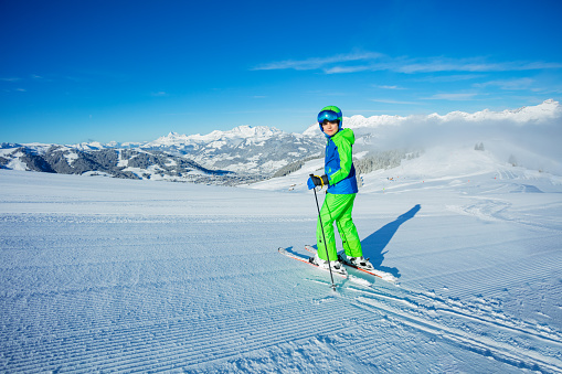 Preteen boy stand on alpine ski look back and smile, about to go down fresh skiing track with mountains peak on background on sunny morning