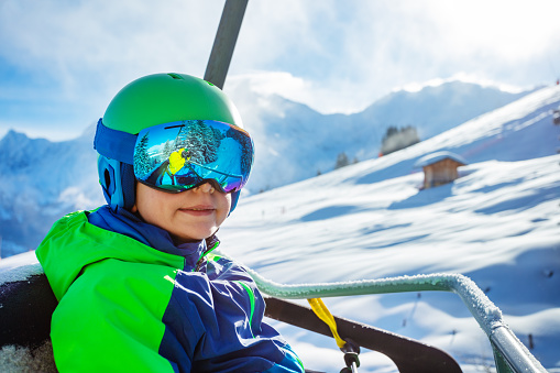 Close face portrait of a young boy sit in sport outfit wear helmet mask and ski going up on chairlift over Alpine background
