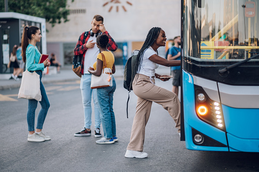 Multiracial passengers waiting at a bus stop and talking cheerfully. Focus on an african american woman entering the bus. Diverse tourists in new town. Best friends traveling together in a city.