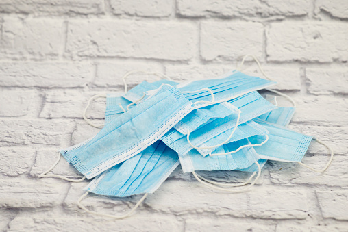 A pile of used blue medical face masks lay in a large pile on a white brick background showing the environmental waste that the covid-19 pandemic is causing.