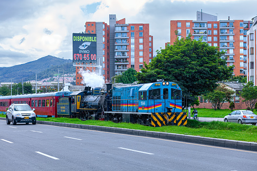 Bogota, Colombia - May 18, 2014: The steam engine pulling the Tourist Train, or in Spanish, 'Tren Turistico' is in trouble.  Rare photo of a diesel engine pulling the steam engine and the train as they return from the Sabana.  Photo shot in the Cedritos area of the capital city of Bogota, Colombia, in South America. On either side of the track is the Carrera Novena; some cars can be seen on the Carrera.  In the background are apartment blocks. and the Andes mountains. An advertising hoarding says in Spanish, 'Disponible', translated, it means, 'Available'.  Photo shot in the horizontal format, in the late evening light; the sky is overcast. Copy space.