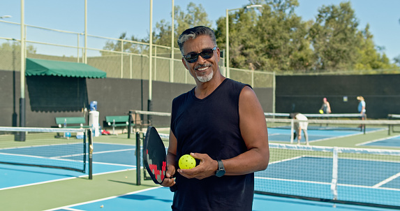 Portrait of an energetic Latino man in his early sixties posing for the camera at an established complex of pickleball courts.