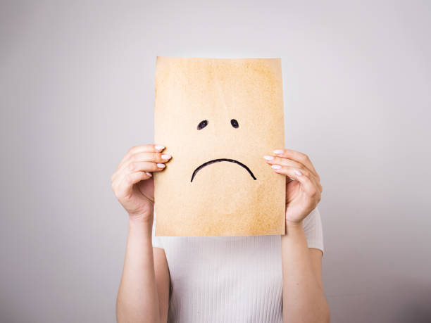 young woman hides her face behind a sheet of paper with a sad smiley face drawn on a gray background, young woman hides her face behind a sheet of paper with a sad smiley face drawn on a gray background, selective focus seasonal affective disorder stock pictures, royalty-free photos & images