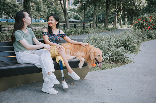 Disable Asian Indonesian woman with prosthetic leg talking to her friend sitting on park bench with her dog