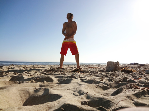 Man wearing swimshorts brushes sand off hands as he stands on beach in Santa Barbara, California.