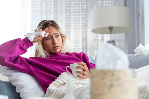 Sick woman with headache sitting under the blanket. Sick woman with seasonal infections, flu, allergy lying in bed. Sick woman covered with a blanket lying in bed with high fever, Coronavirus symptoms