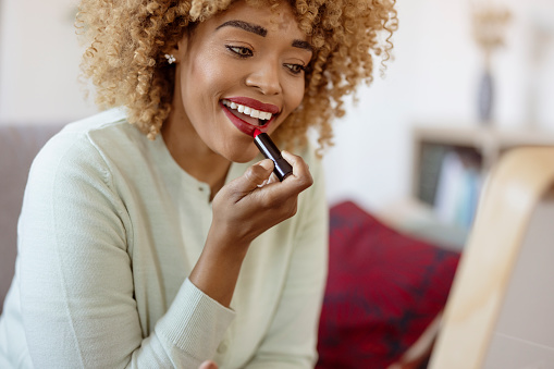 Happy curly african american woman putting lipstick on at home