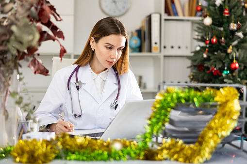 Portrait of successful female doctor physician working with paperwork and laptop in office at Christmas