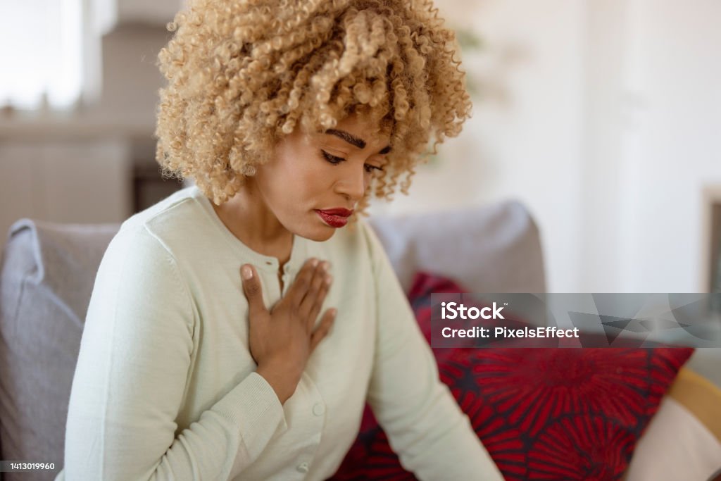 Let's just hope this isn't serious African american woman feeling sick and holding her chest in pain while sitting on the sofa in the living room One Woman Only Stock Photo