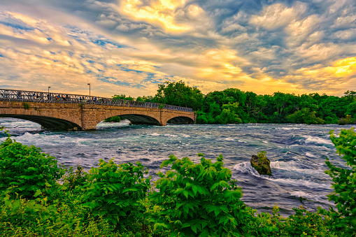 Bridge across rapids splash Niagara falls river with green summer flowers in front over colorful clouds sky between USA and Canada