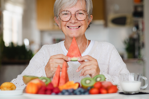 Beautiful happy senior woman holding a slice of watermelon while having breakfast at home with fresh seasonal fruit, milk and cupcake, healthy eating concept