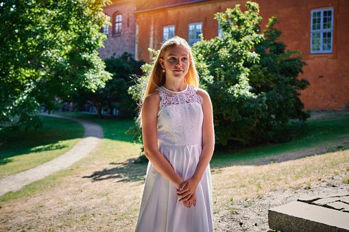 Fourteen year old girl in her official white confirmation dress standing outdoors in a garden