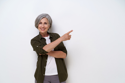 Pointing sideways grey haired mature businesswoman with hands folded looking straight in camera wearing green shirt and white t-shirt isolated on white background. Mature people beauty concept.