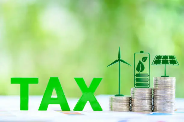 Photo of Clean, renewable energy or electricity production tax credits and incentives, financial concept : Green energy symbols atop coin stack e.g solar panel, wind turbine, fuel cell battery and the word TAX