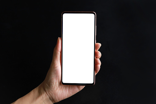 Cropped shot of a woman's hand holding up a smartphone with blank white screen against black background. Lifestyle and technology. Smartphone with blank screen for design mockup.