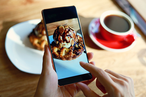 Cropped shot of a young woman photographing pastry and coffee on the table in a cafe with smartphone