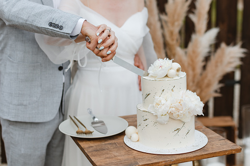 Bride and groom cutting stylish wedding cake at wedding in outdoor. Wedding couple holding knife and cutting together wedding cake decorated with flowers