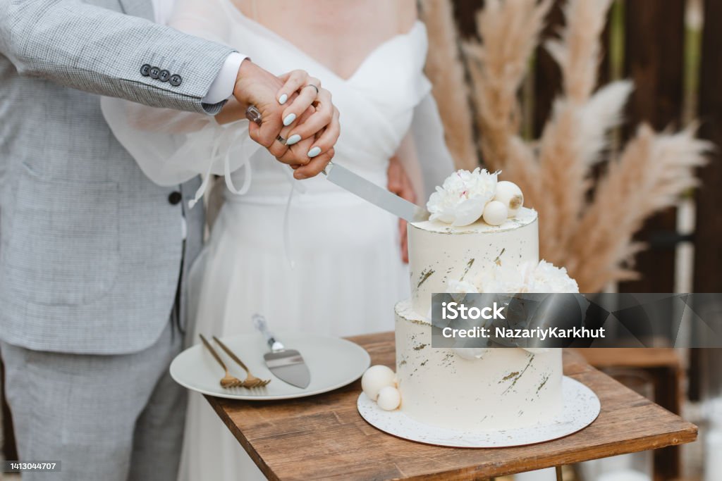 Bride and a groom is cutting their beautiful wedding cake on wedding banquet. Hands cut the cake with delicate white flowers. Bride and groom cutting stylish wedding cake at wedding in outdoor. Wedding couple holding knife and cutting together wedding cake decorated with flowers Wedding Cake Stock Photo