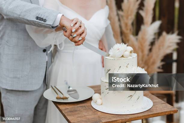 Bride And A Groom Is Cutting Their Beautiful Wedding Cake On Wedding Banquet Hands Cut The Cake With Delicate White Flowers Stock Photo - Download Image Now