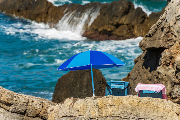 Blue beach umbrella and two deck chairs on the rocky coast in Liguria, Italy, Europe. Tellaro village, tourist resort on the coast of Gulf of La Spezia or Gulf of Poets (Golfo dei Poeti), Lerici municipality, Mediterranean sea.