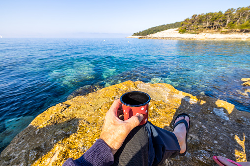 Man holding red coffee cup at rocky beach by sea on sunny day