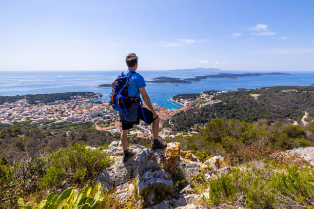 man with backpack standing on rock and enjoying view of town from top of mountain - sky sea town looking at view imagens e fotografias de stock