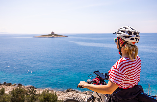 Female biker wearing helmet holding bicycle while looking at beautiful calm blue sea on sunny day