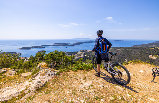 Male cyclist with backpack and bike looking at beautiful coastal town from mountain top on sunny day