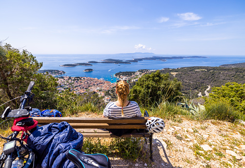 Female biker sitting on wooden bench while looking at beautiful coastal town from mountain top on sunny day