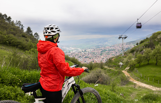 Female biker wearing helmet and red jacket with bicycle watching at ski lift on grassy mountain slope in countryside