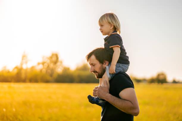 istock Father carries his son on shoulders while walking on grass field against sky 1413051728