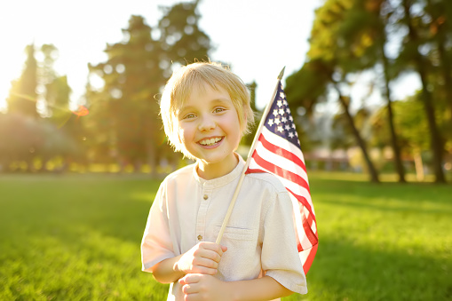 Cute little boy celebrating of July, 4 Independence Day of USA in park at sunny summer sunset. Child holding american flag symbol of United States. Pride and patriotism concept.