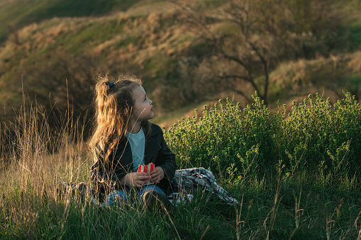 The family spends time on a picnic outside the city, a little girl with curly hair sits on the ground in the rays of the setting sun