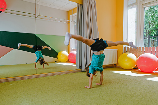 Little girl doing handstand with twine in sport gym. Preschooler doing acrobatic cartwheel on the mat. Health concept.