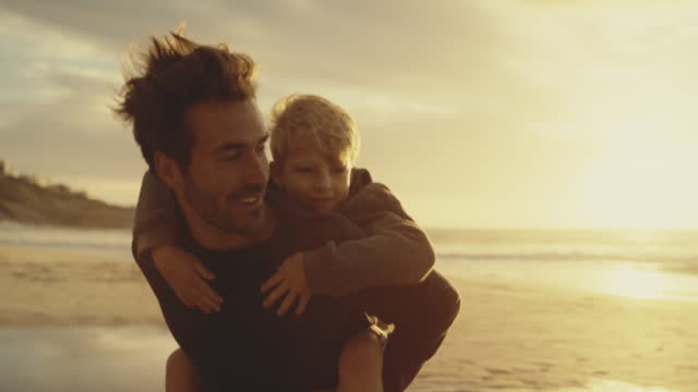 Excited and happy, cute little boy riding piggyback on his dad during a sunset walk at the beach. Happy father and son having fun and spending time together by the sea while on summer vacation