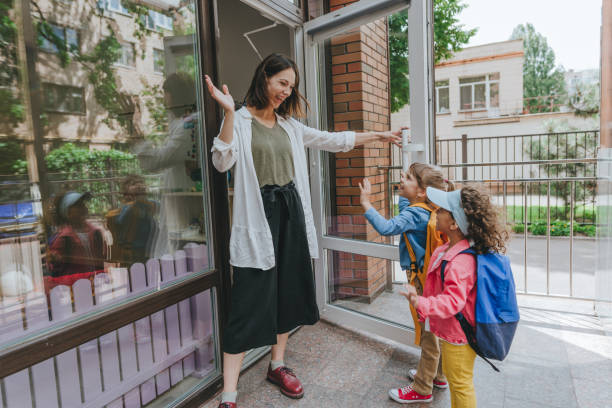 Happy smiling woman teacher greeting elementary school students Happy smiling woman teacher greeting elementary school students at first day at school standing near the open door outside. Selective focus. kid waves stock pictures, royalty-free photos & images