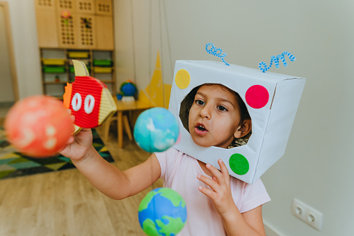 Funny little girl wearing handmade helmet playing with paper spaceship learning Solar system planets models at home or kindergarten. Education science concept. Selective focus.