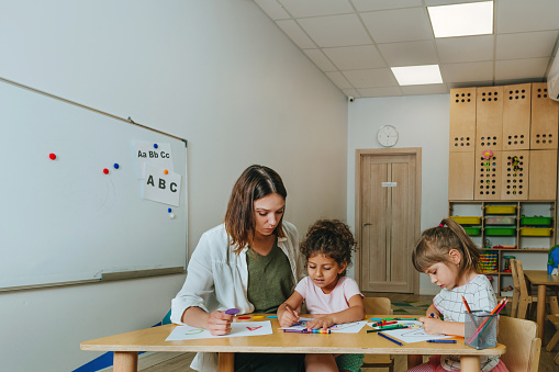 English lesson at elementary school or kindergarten. Students learning alphabet and colors with teacher coloring the letter A sitting in the classroom. Selective focus.