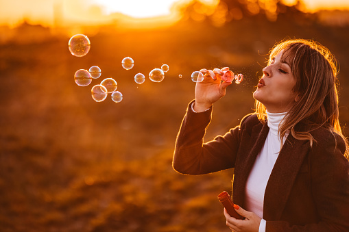 Profile of a beautiful young woman blowing bubbles in the park at sunset during autumn.